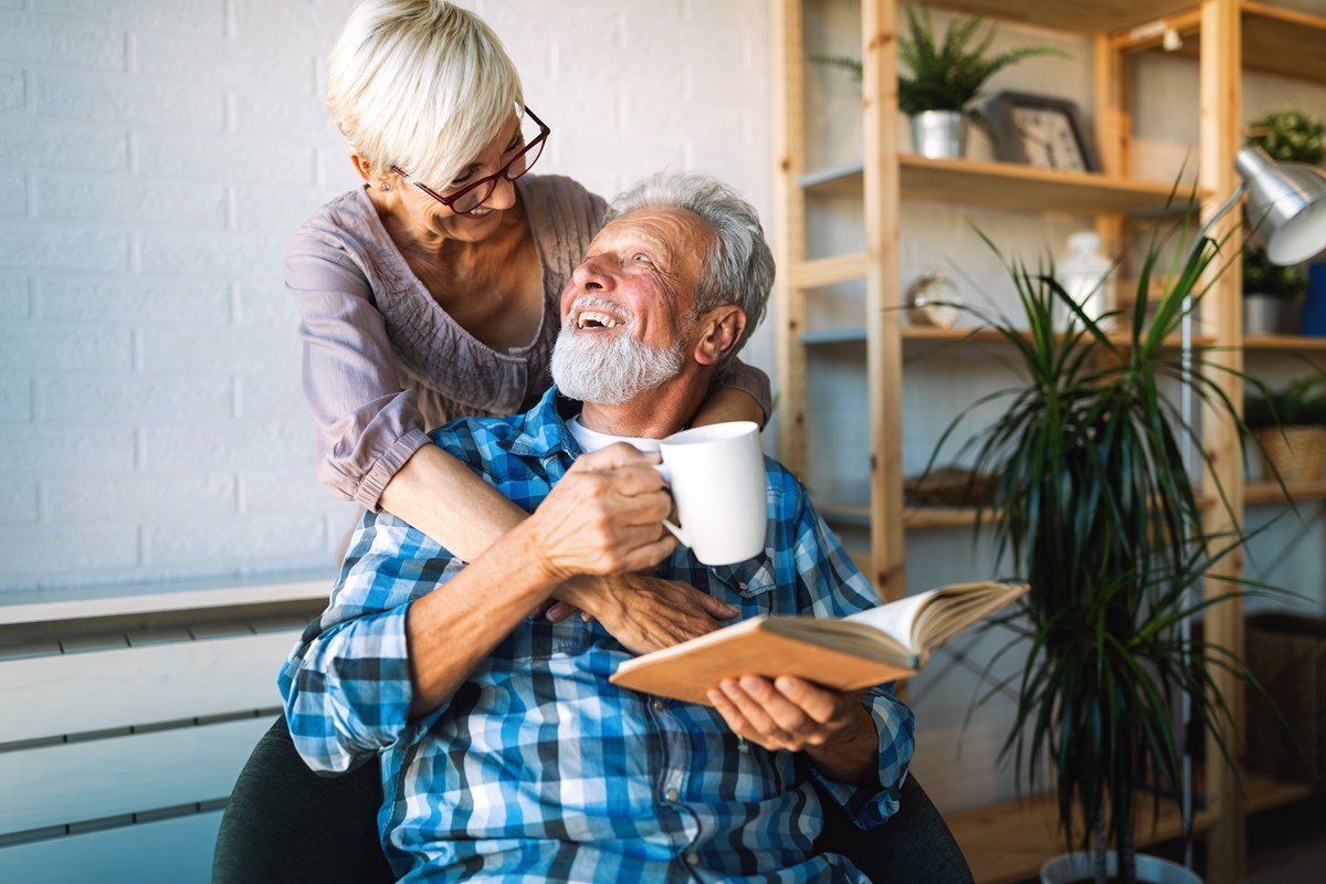 A man sitting on a chair while reading a book. A woman has her arms around him..